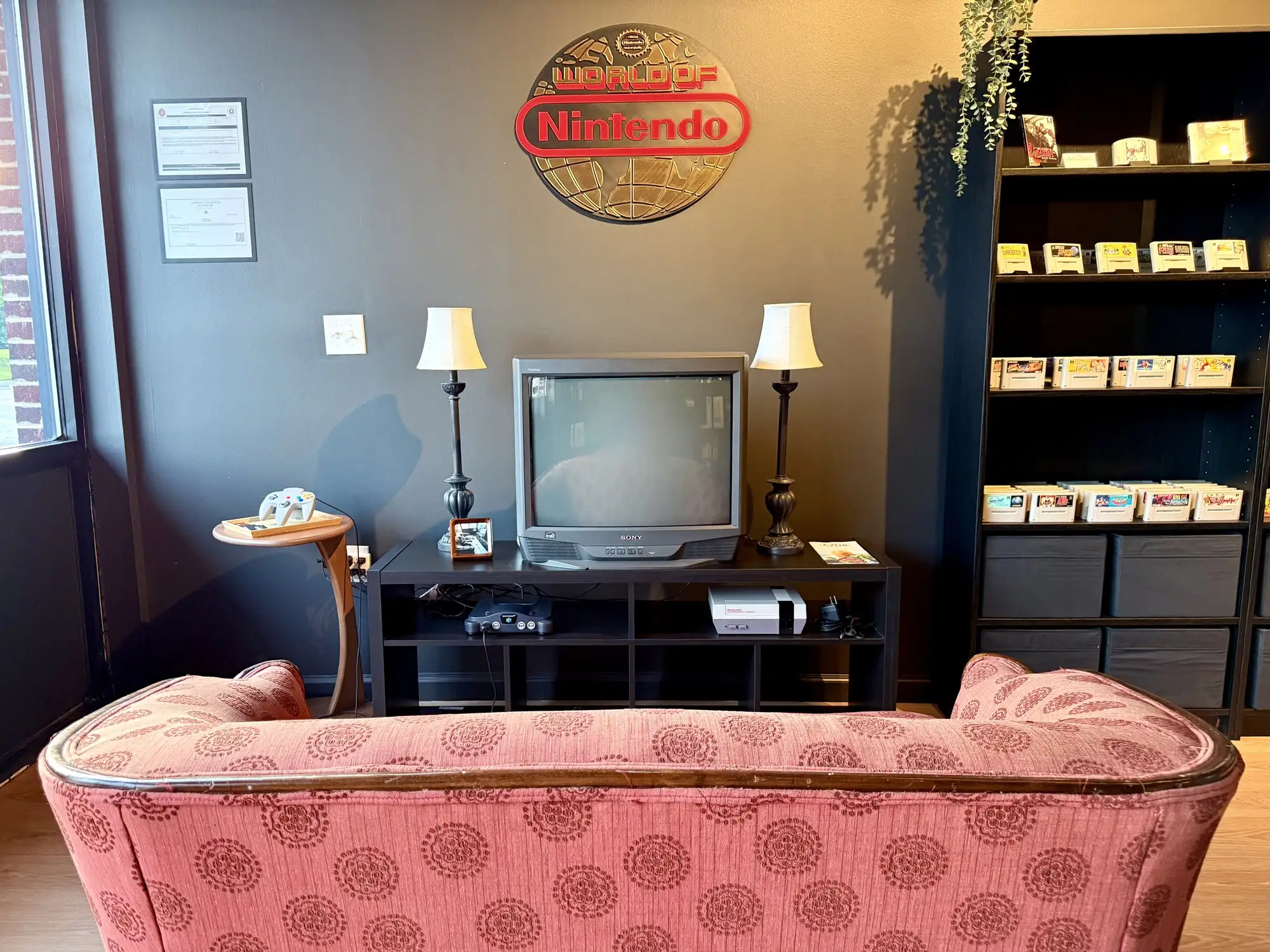 The play corner at NOSTOS in Duluth, GA: a Sony CRT television on a low shelf with a Nintendo 64 and an NES beneath it, a World of Nintendo sign on the wall above, and a vintage sofa facing the screen